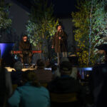 Muzyczny występ na scenie, dwóch artystów; jeden gra na keyboardzie, drugi śpiewa. W tle zielone drzewa, scena oświetlona. Publiczność ogląda koncert siedząc na krzesłach.