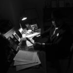 Czarno-białe zdjęcie przedstawia osobę w kapeluszu, siedzącą przy biurku otoczonym zdjęciami. Na biurku leży lampa i papiery. Tło jest ciemne, a oświetlenie tworzy nostalgiczny klimat.