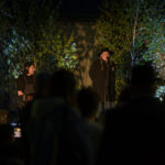 Dwóch wykonawców na scenie, jeden gra na klawiaturze, drugi śpiewa. Tło oświetlone zielonym światłem, dekoracja z drzewami. Wieczorna atmosfera z publicznością.