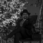 Mężczyzna w kapeluszu siedzi na krześle, ubrany formalnie. W tle liście drzewa, scena oświetlona reflektorami. Obraz czarno-biały.