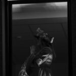 Kobieta w kapeluszu ozdobionym piórami stoi przy oknie. Ubrana w elegancką suknię. Tło jest jasne, wnętrze z sufitem panelowym.