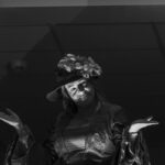 Kobieta w kapeluszu z kwiatami i stroju historycznym, unosi ręce w geście niepewności. Zdjęcie czarno-białe, ciemne tło.świetlenie skupione na twarzy.