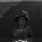 Kobieta w historycznym stroju z kapeluszem z piórami stoi w cieniu. Tło ciemne, sufit jasno oświetlony. Ujęcie czarno-białe.