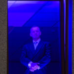 Zdjęcie przedstawia osobę w ciemnym garniturze stojącą za oknem. Tło jest ciemne z delikatnym fioletowym oświetleniem. Ramka okna brązowa, ściana szara.