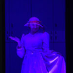 Obraz przedstawia witraż z postacią ubraną w elegancką suknię i kapelusz. Dominują kolory fioletowy i różowy. Tło jest ciemne, co podkreśla kontury postaci.