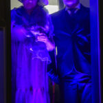 Na zdjęciu są dwie osoby w strojach z epoki, oświetlone na fioletowo. Tło jest ciemne, z zarysem okna. Wyraziste, retro nakrycia głowy.