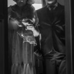 Na czarno-białym zdjęciu starsza para w eleganckich ubraniach stoi na tle okna. Kobieta ma kapelusz i futrzasty szal. Mężczyzna w kapeluszu i garniturze.