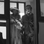 Na czarno-białym zdjęciu dwoje ludzi stoi za szybą. Kobieta w kapeluszu i futerku, mężczyzna w garniturze i cylindrze. W tle widać sufit z paneli.