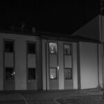 Budynek nocą z jasno oświetlonymi oknami. W oknach widać sylwetki ludzi. Na ciemnym tle widać zegar i rowery.