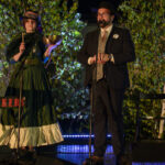 Dwóch ludzi na scenie w eleganckim stroju, na tle zielonych liści. Kobieta w zielonej sukni trzyma misę. Obok mężczyzna w kapeluszu przy mikrofonie.