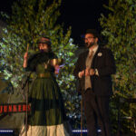 Para artystów na scenie, kobieta w zielonej sukni trzyma miskę. Mężczyzna w garniturze obok mikrofonu. W tle oświetlone drzewa.