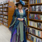 Osoba w eleganckiej sukni z kapeluszem znajduje się w bibliotece. Suknia w zieleni i fioletach harmonizuje z książkami na półkach. Tło stanowią regały z książkami oznaczonymi kategoriami.