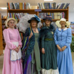 Cztery kobiety w strojach retro pozują przed półką z książkami. Mają kolorowe suknie i kapelusze, jedna trzyma parasolkę. Tło wypełnione jest książkami.