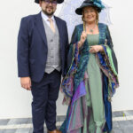 Mężczyzna w granatowym garniturze i kobieta w barwnej sukni z parasolką. Stoją na tle jasnej ściany z szarymi płytkami.