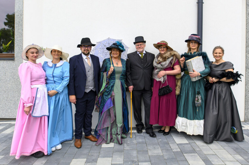 Grupa ludzi w strojach vintage pozuje na tle białej ściany. Kobiety w kolorowych sukniach, mężczyźni w kapeluszach i garniturach. Na ziemi szare płytki.