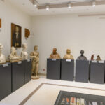 Na zdjęciu widzimy galerię popiersi i rzeźb w jasnym pomieszczeniu. Popiersia stoją na szarych postumentach, a tło jest kremowe. Nad popiersiami wisi obraz portretowy.