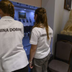 Dwie osoby w białych koszulkach GMINA DOBRE stoją w pomieszczeniu galeryjnym. Obok znajduje się rzeźba, a w tle widoczne są obrazy. Atmosfera jest elegancka i artystyczna.