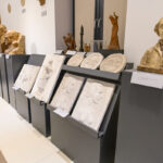 Galeria z popiersiami, płaskorzeźbami i rzeźbami na czarnych postumentach. Jasne ściany, kontrastujące ciemne podstawy. Rzeźby w ciepłych odcieniach brązu i bieli.