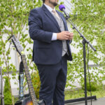 Mężczyzna w garniturze i kapeluszu stoi przy mikrofonie z fioletową osłoną. Obok niego znajduje się gitara. W tle zielone drzewa.