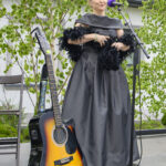 Na zdjęciu kobieta w szarej sukni śpiewa do mikrofonu na tle zieleni. Obok stoi gitara o drewnianej fakturze. Tło stanowi bujna roślinność.