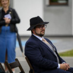 Mężczyzna w ciemnym garniturze i kapeluszu siedzi na zewnątrz. Za nim stoją osoby w jasnym tle budynku. Atmosfera formalna.