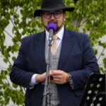 Mężczyzna w kapeluszu i okularach przemawia do mikrofonu. Ubrany elegancko w granatową marynarkę na tle zielonych liści.