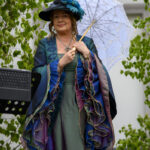 Kobieta w kapeluszu z piórami trzyma jasną parasolkę. Nosi kolorową, falistą suknię. W tle zielone liście i biały mur.