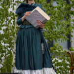 Kobieta w kapeluszu trzyma książkę, ubrana w zieloną suknię z białą falbaną. W tle zielone liście drzew.