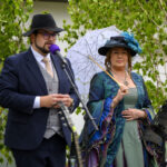 Na zdjęciu są mężczyzna w garniturze i kobieta w eleganckiej sukni z parasolką. Tło stanowią zielone krzewy. Oboje stoją przy mikrofonie i mównicy.