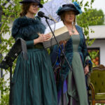 Dwie kobiety w strojach historycznych w odcieniach zieleni i niebieskiego stoją przy mikrofonie na tle zieleni. Jedna trzyma parasolkę, druga księgę. Tło tworzy natura, z dużą ilością zielonych liści.