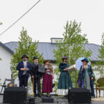 Na zdjęciu pięć osób w strojach retro na zewnątrz. W tle zielone drzewa i biały budynek. Scena romantyczna z parasolką.