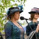 Dwie kobiety w stylowych kapeluszach śpiewają przy mikrofonach. Ubrania w odcieniach zieleni i turkusu. Tło z liściastymi gałęziami.