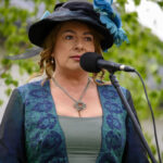 Kobieta w eleganckim stroju przemawia do mikrofonu. Ubrana w zielono-niebieską suknię i czarny kapelusz z niebieskim piórem. W tle zielone liście.