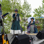 Na scenie na zewnątrz występują trzy osoby w historycznych strojach. Jest dużo zieleni, między innymi drzewa w tle. Obok sceny stoi gitara i mikrofony.