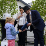 Dziewczynka w różowych spodniach otrzymuje prezent od mężczyzny w kapeluszu. Kobieta w białej marynarce stoi obok. W tle są drzewa.