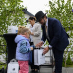 Na zdjęciu mężczyzna w kapeluszu wręcza torbę dziewczynce. W tle zielone drzewa i kobieta w białej marynarce. Event na zewnątrz.