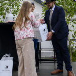 Na zdjęciu osoba w kapeluszu wręcza torbę dziewczynce w beżowej odzieży. Tło jest zielone z drzewami. Obie postacie są uśmiechnięte.