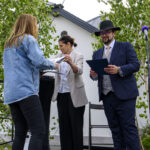 Trzy osoby na scenie, jedna wręcza dokument, druga trzyma teczkę. W tle drzewka i jasny budynek.