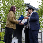 Młoda kobieta odbiera dyplom od mężczyzny w kapeluszu. Drzewka w tle, białe torby na ziemi. Scena na zewnątrz, elegancka atmosfera.