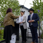 Na zdjęciu są trzy osoby na scenie, w tym jedna wręcza torbę drugiej. Tło to drzewa i budynek. Kolory są stonowane, dominują brązy i zielenie.