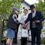 Na scenie mężczyzna w kapeluszu i garniturze wręcza nagrodę chłopcu w kamizelce. Obok kobieta trzyma białą torbę. Tło to zielone drzewka i biały budynek.