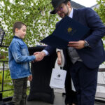 Dwóch mężczyzn w eleganckiej odzieży wymienia się uściskiem dłoni. Jeden z nich trzyma granatową teczkę i biały worek. W tle zielone drzewa.