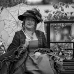 Kobieta w eleganckiej sukni z parasolką siedzi na zewnątrz obok retro telefonu. Tło to drzewa i ogrodzenie. Zdjęcie jest czarno-białe.