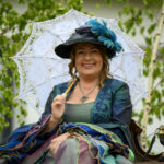 Osoba w stylowym, zielonym stroju retro siedzi, trzymając białą parasolkę. Na głowie ma czarny kapelusz z niebieskimi piórami. W tle drzewa i starodawny telefon.