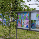 Wystawa rysunków dziecięcych na zewnętrznych tablicach. Kolorowe prace, zielone drzewa w tle. Użyte lampki tworzą przyjemną atmosferę.