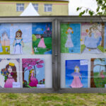 Tablica z dziecięcymi rysunkami przedstawiającymi księżniczki i zamki. Rysunki w jasnych kolorach. W tle budynek i drzewa.