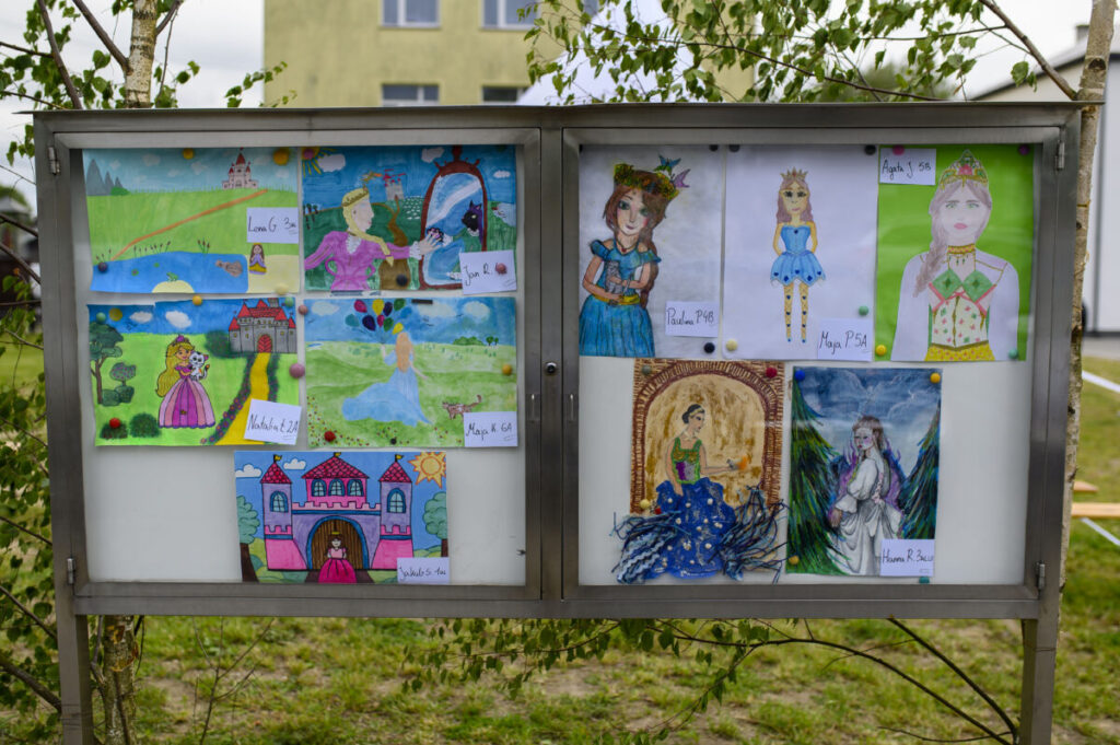 W gablotce wiszą kolorowe dziecięce rysunki postaci i zamków. Obrazki na białym tle, otoczone zielenią drzew. Tło to jasny budynek.