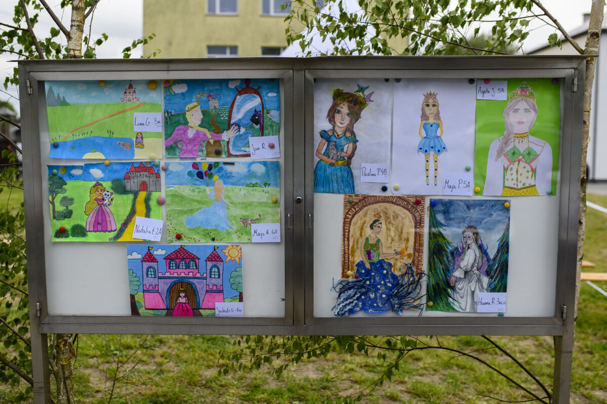 W gablotce wiszą kolorowe dziecięce rysunki postaci i zamków. Obrazki na białym tle, otoczone zielenią drzew. Tło to jasny budynek.