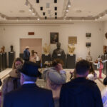 Na obrazie widoczna jest sala muzealna z rzeźbami i popiersiami. Ściany są jasne, a wystawa ma eleganckie oświetlenie. Kilka osób ogląda eksponaty w tle.