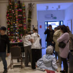 Wnętrze galerii z kolorowymi palmami wielkanocnymi na ścianie. Ludzie w różnym wieku zwiedzają ekspozycję rzeźb. Jasne, nowoczesne wnętrze.